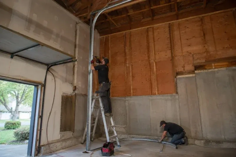 One person on a stepladder working beside a tall curved garage-door track while another kneels beside loose track sections in an unfinished garage. — High-Lift Garage Door Conversion, Toronto